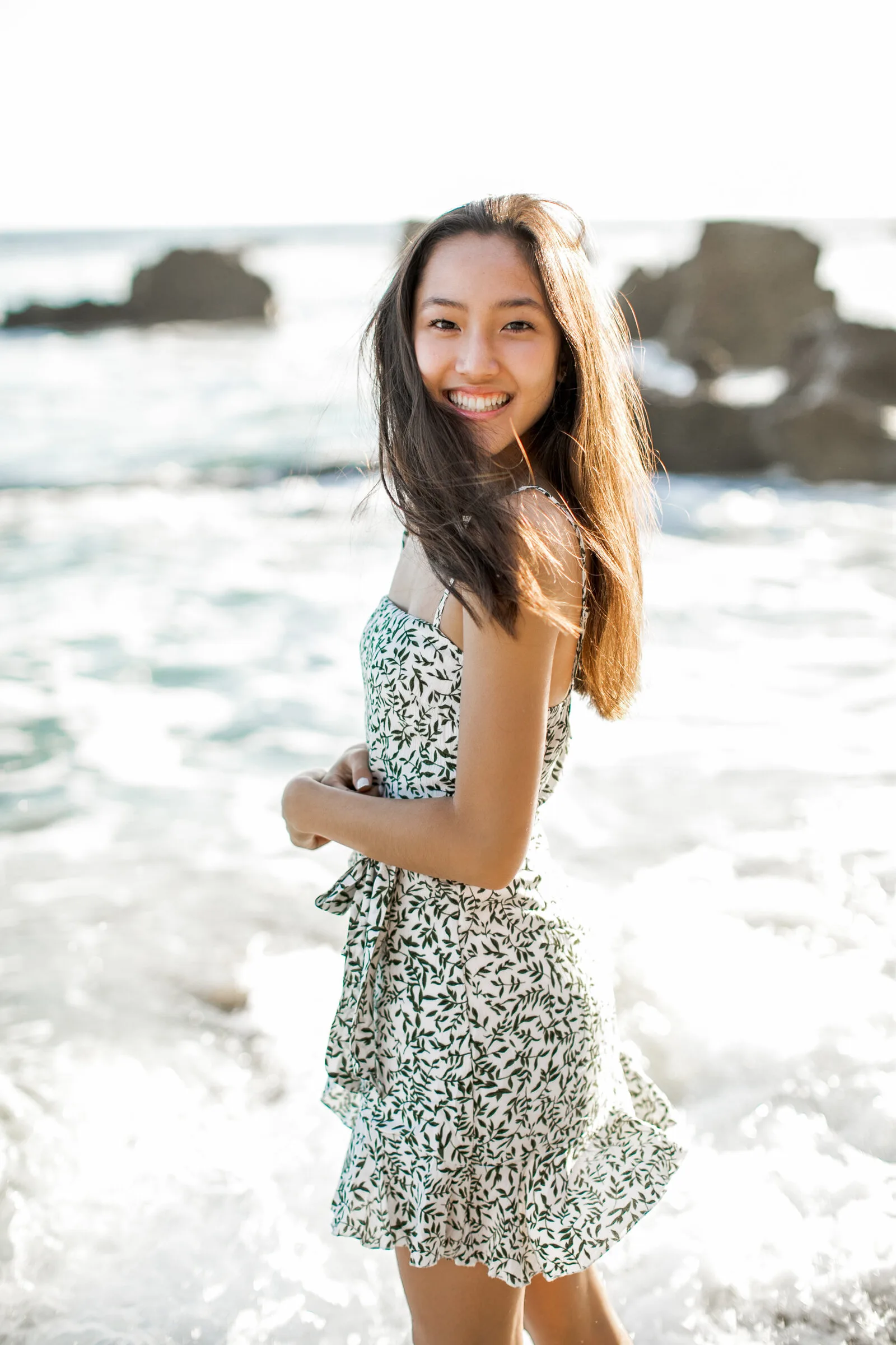 A girl in a leaf-print dress smiling over her shoulder with surf behind