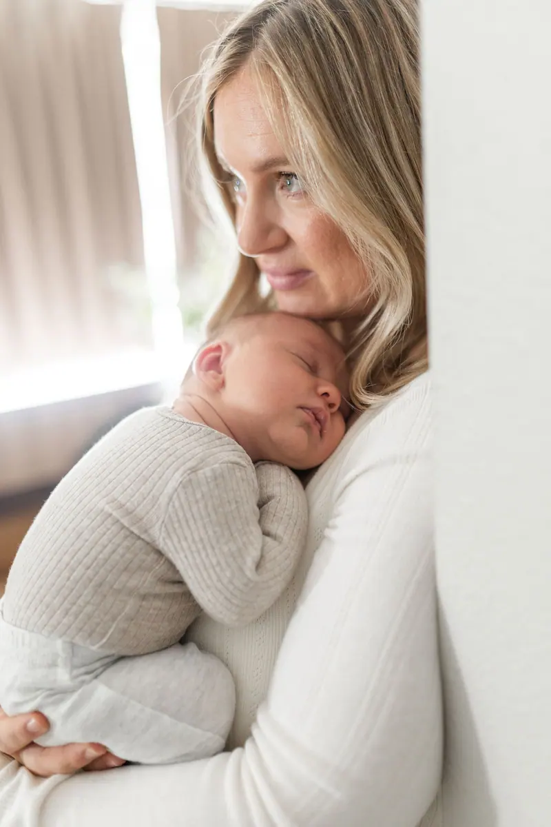 A mother holding her sleeping newborn against her chest by a window