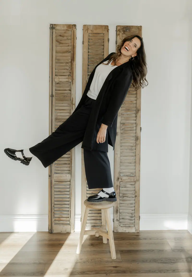 A woman in a long black coat laughing and kicking one leg out on a stool
