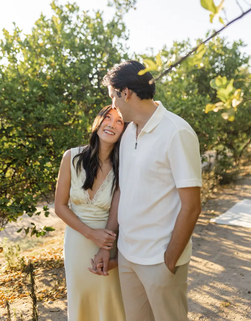 A man kissing his fiancée's forehead in a sunlit grove