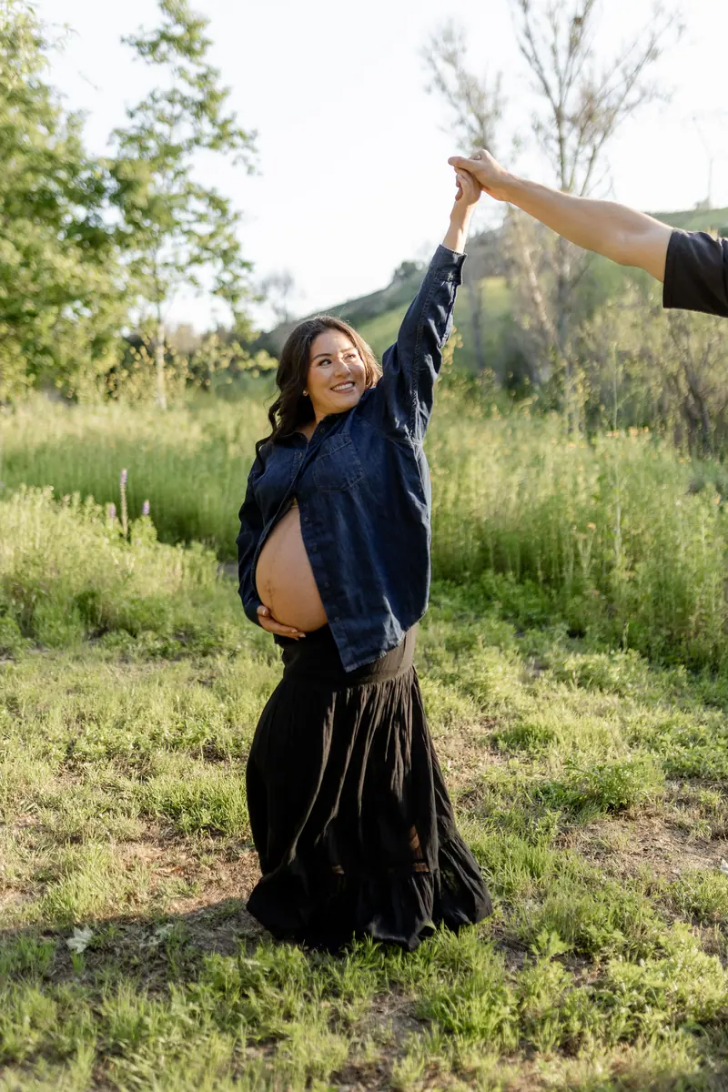 An expecting mother laughing in a green field, reaching for her partner's hand