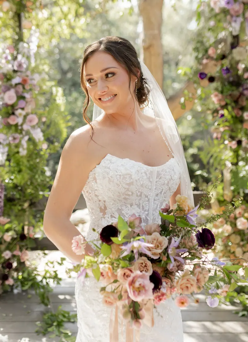 A bride smiling over her shoulder with a pastel bouquet by the flower arch
