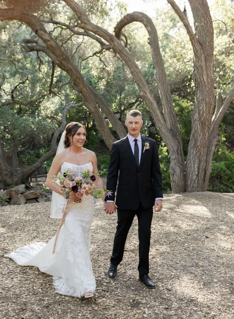 A bride and groom holding hands beneath sprawling oak branches