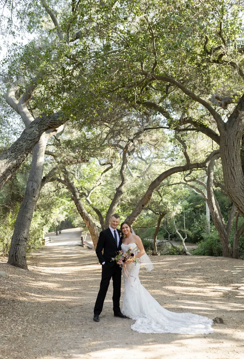 A bride and groom standing on a dirt path beneath arching oaks